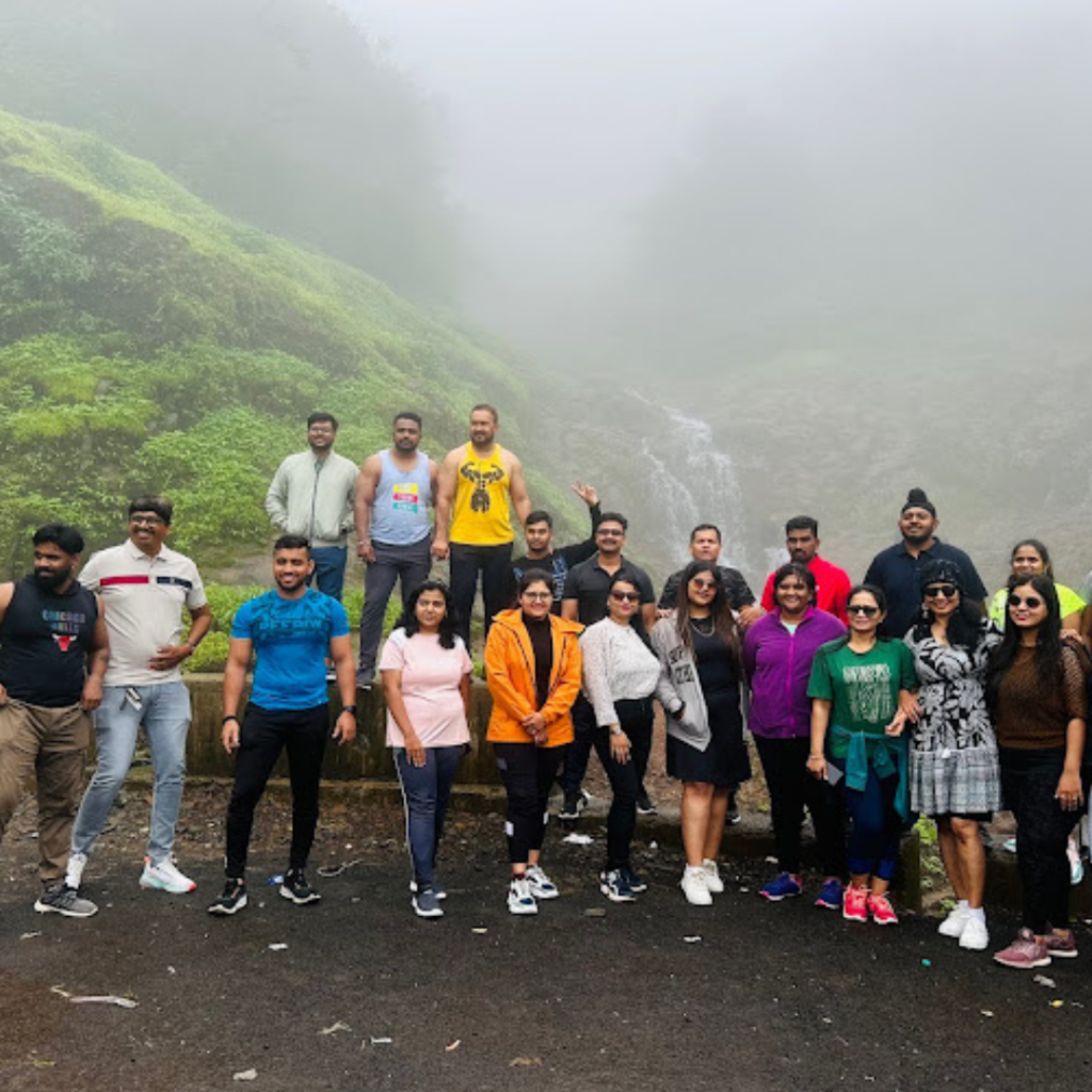 "Group of fitness enthusiasts posing for a photo during an outdoor fitness trip with a scenic waterfall and mist-covered green hills in the background."