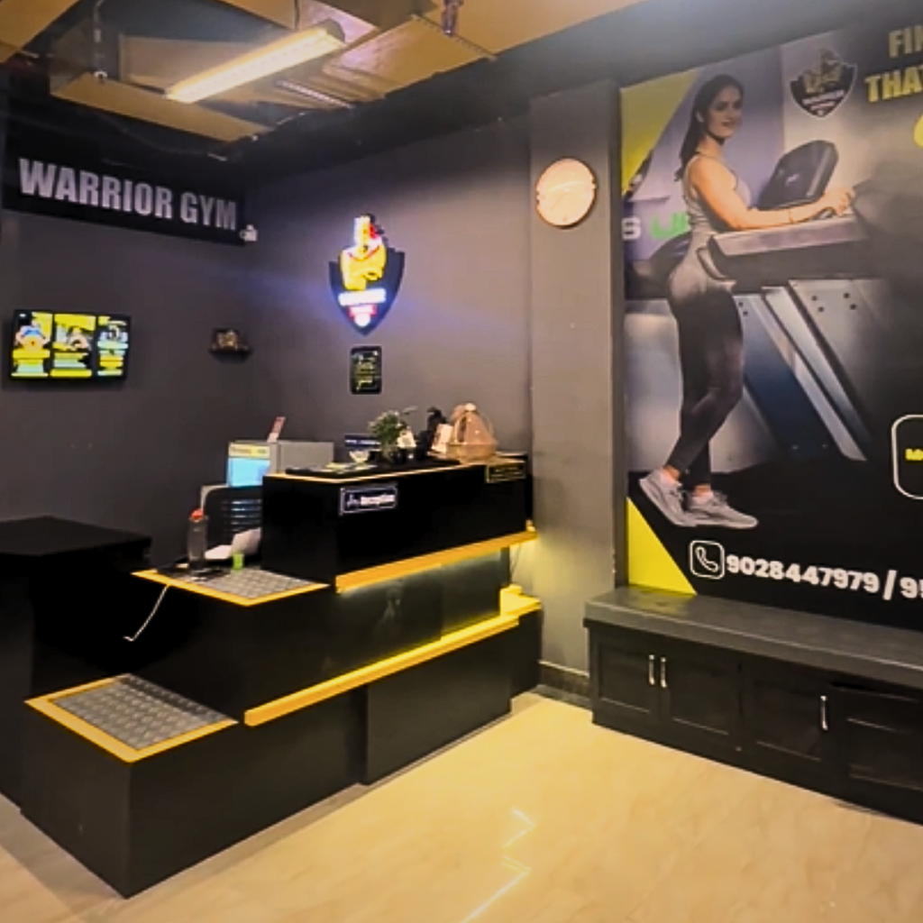 "Modern gym reception area with a welcome desk, Warrior Gym branding, illuminated logo, and fitness-themed wall graphics creating a professional entrance."