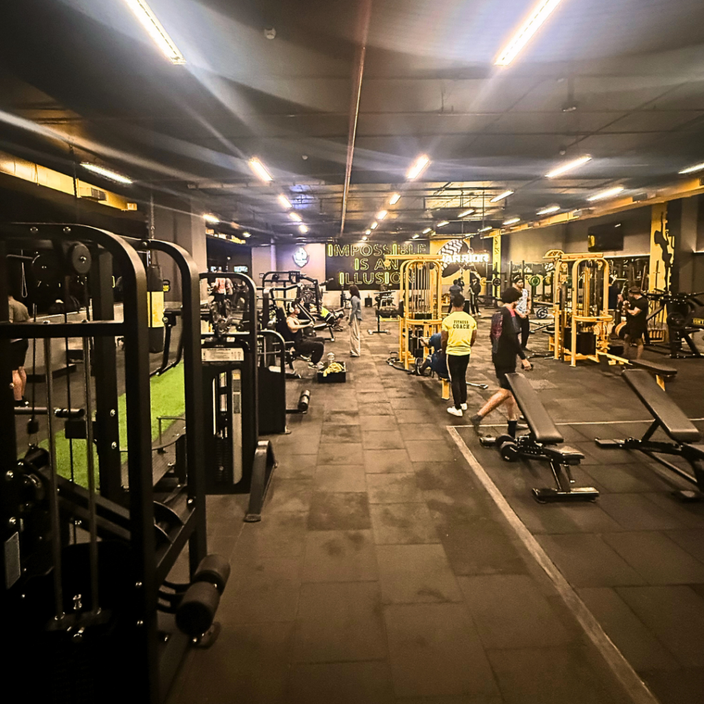 "Spacious modern gym with strength training machines, free weights, workout benches, and members exercising in a well-equipped fitness center."