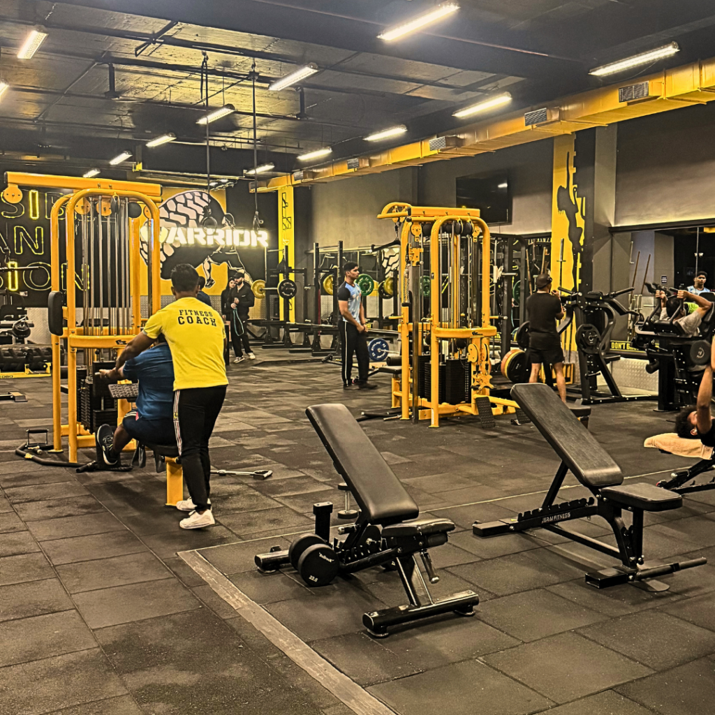 A spacious modern gym with yellow-and-black strength machines, adjustable benches, free weights, and rubber flooring.